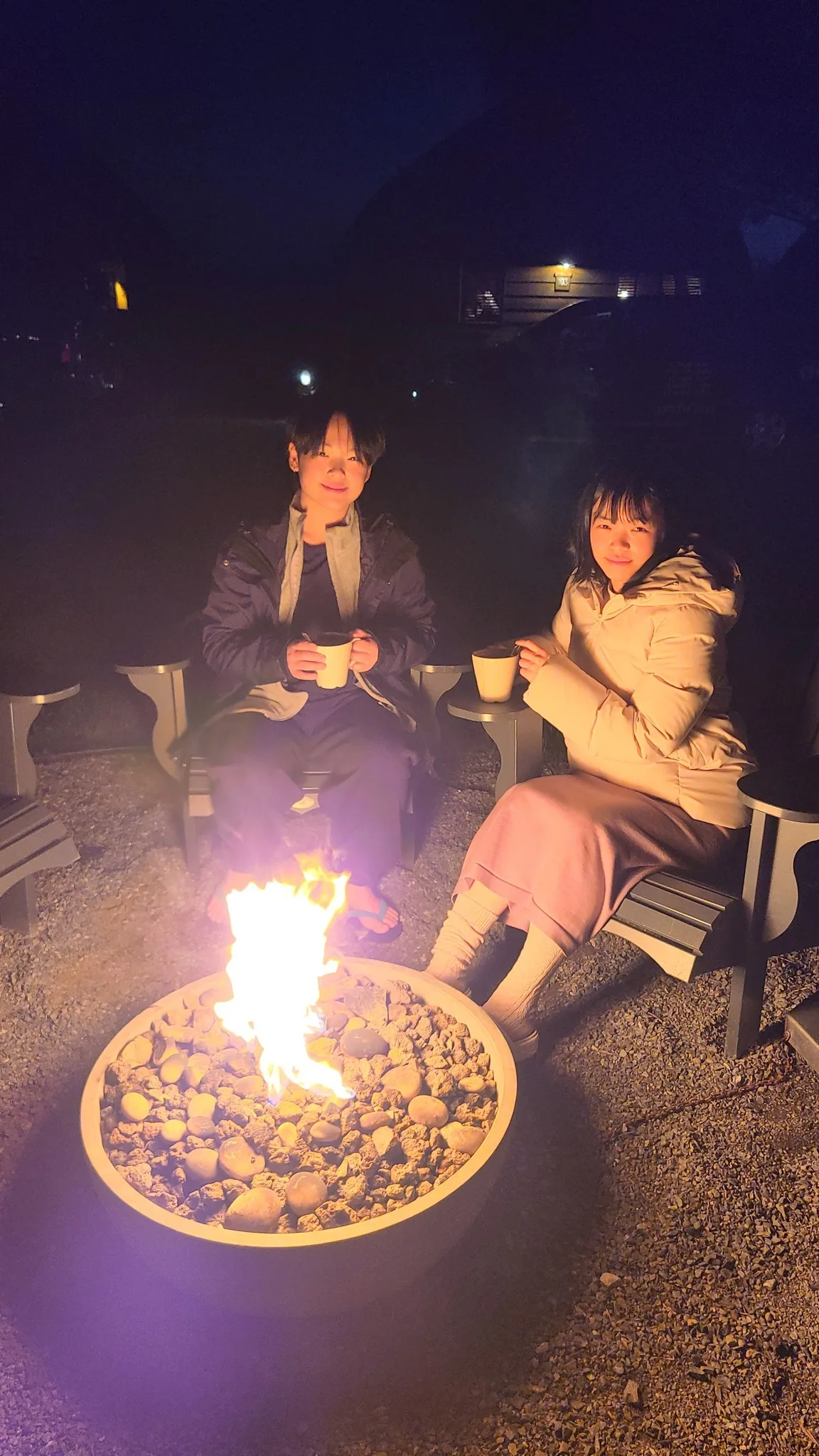 Two family members sitting by a campfire with warm drinks outdoors at night